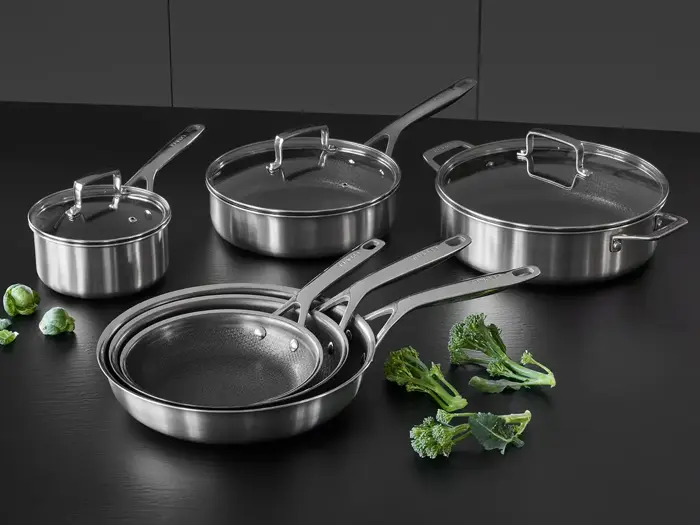 Et sæt gryder og pander i rustfrit stål med låg på en sort overflade, med et par stykker broccoli og rosenkål i nærheden.