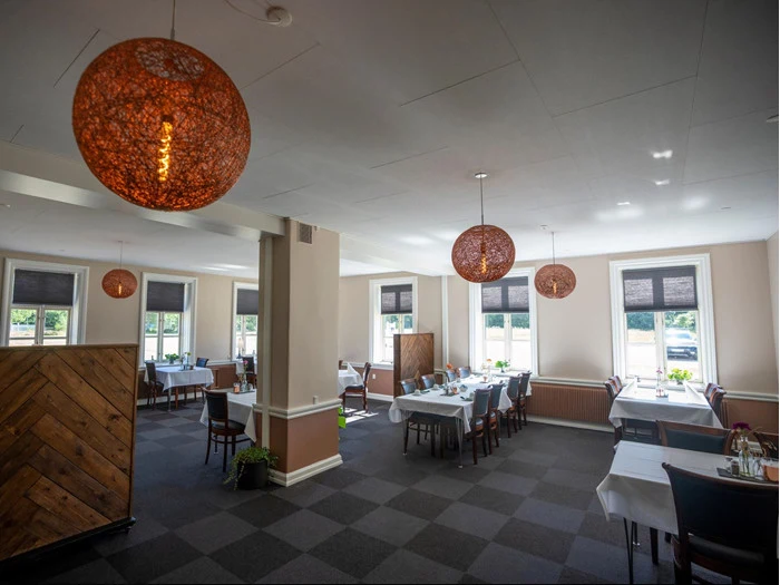 En rummelig, solbeskinnet restaurant med store vinduer, mørke stole, hvide duge og runde orange hængelamper.