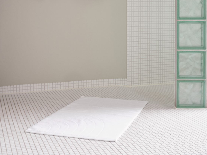 Et minimalistisk badeværelse med hvide kvadratiske fliser, en hvid bademåtte på gulvet og en del af væggen lavet af glasblokke.