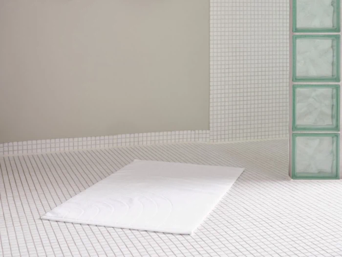 Et minimalistisk badeværelse med hvide kvadratiske fliser, en hvid bademåtte på gulvet og en del af væggen lavet af glasblokke.