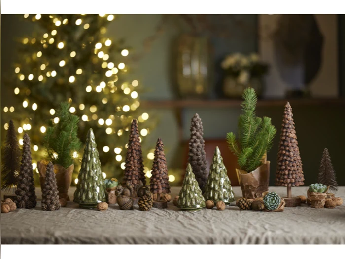 En samling små, dekorative juletræer lavet af kogler og grønt står på et bord med et sløret, oplyst juletræ i baggrunden.
