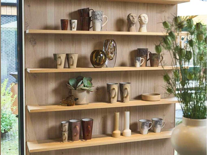 Træhylder med forskellige keramikkrus, kopper, vaser, dekorative buster, stearinlys og en sukkulent i potte med planter, der kan ses gennem vinduet ved siden af hylderne.