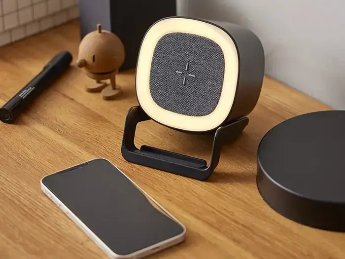 En lille, moderne trådløs højttaler med et glødende lys står på et træbord ved siden af en smartphone og andet skrivebordstilbehør.