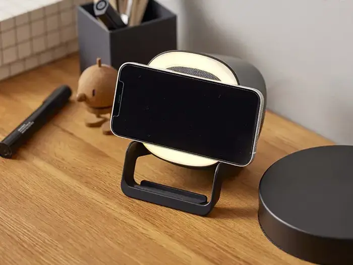 En smartphone hviler vandret på et trådløst opladningsstativ på et træbord med kontorartikler og en lille figur i baggrunden.