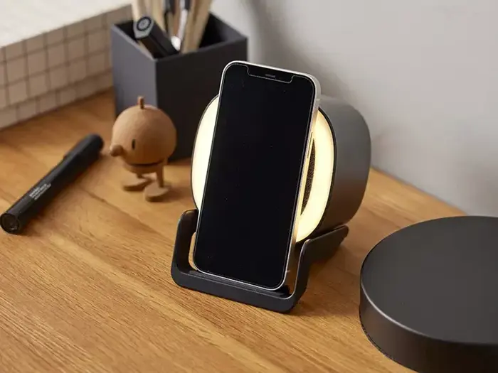 En smartphone er placeret på et trådløst opladningsstativ på et træbord, omgivet af papirvarer og en lille dekorativ figur.