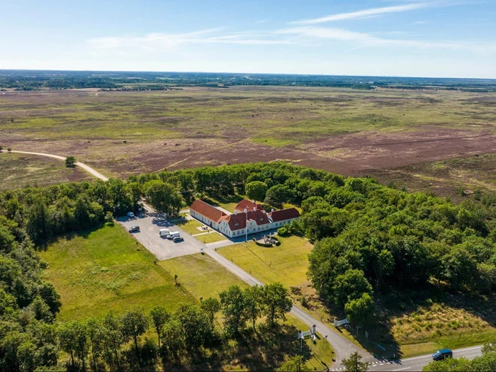 Luftfoto af en stor hvid bygning med rødt tag omgivet af træer i et stort, åbent landskab under en klar blå himmel.