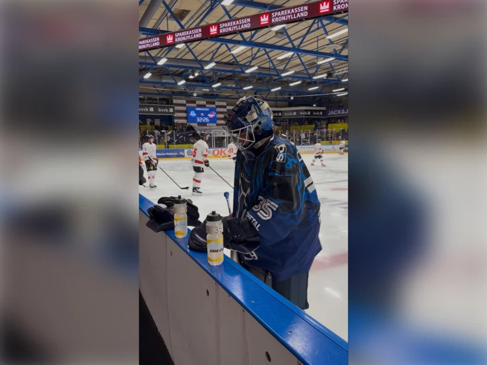 En ishockeymålmand i blåt udstyr står ved banden med vandflasker; i baggrunden træner spillere i hvidt og rødt på isen.