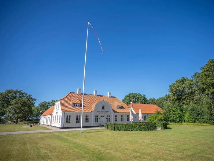 Et stort hvidt hus med rødt tegltag ligger på en velplejet græsplæne under en klar blå himmel med en høj, tom flagstang foran og træer i baggrunden.
