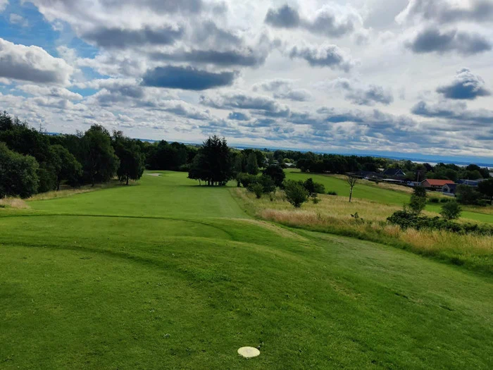 En grøn golfbane med træer, buske og en overskyet himmel i baggrunden på en lys, delvis solrig dag.