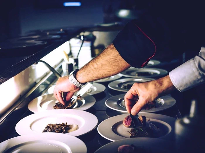En kok anretter gourmetretter og placerer omhyggeligt maden på flere hvide tallerkener, der står på rad og række på en disk i et svagt oplyst køkken.