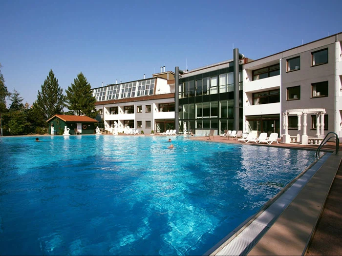 Stor udendørs swimmingpool med et par mennesker i vandet, omgivet af liggestole, foran en moderne hotelbygning i flere etager på en klar, solrig dag.