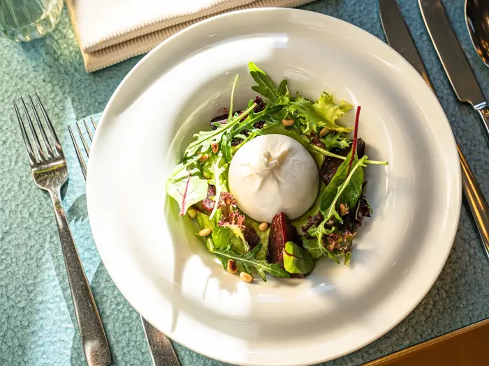 En tallerken med burrataost på en bund af blandet grønt, rødbeder og pinjekerner, sat på et bord med bestik og en foldet serviet i nærheden.