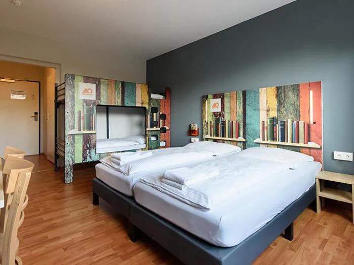 Et hotelværelse med to enkeltsenge, en køjeseng, trægulve, farverige hovedgærder med bogmotiver og minimale træmøbler.