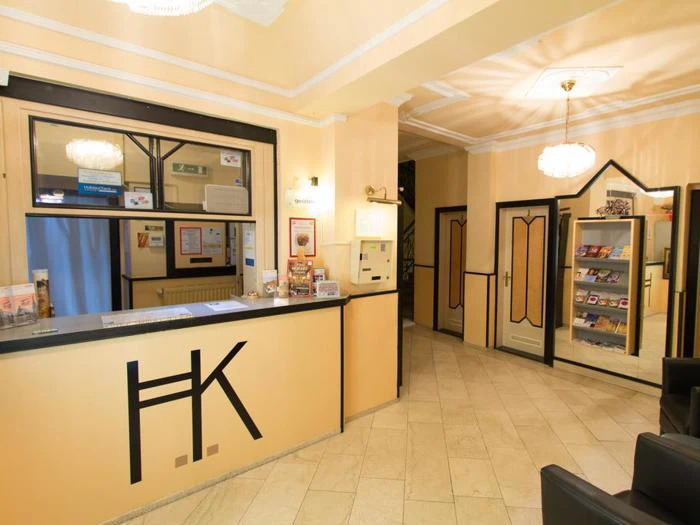 Hotelreception med en skranke mærket HK, brochureholdere, beige vægge, klinkegulv og moderne belysning.