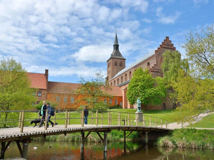 Folk går over en træbro over en dam med en historisk murstenskirke og træer i baggrunden på en delvis overskyet dag.