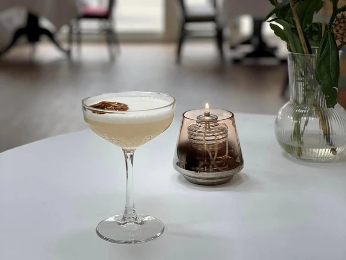 En skummende cocktail pyntet med en tørret citrusskive står på et hvidt bord ved siden af en lille olielampe i glas og en vase med grønne blade.