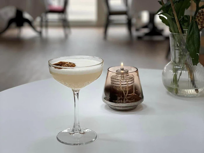 En cocktail med tørret citrus i et coupe-glas står på et hvidt bord ved siden af en tændt olielampe og en vase med grønne blade i et svagt oplyst rum.