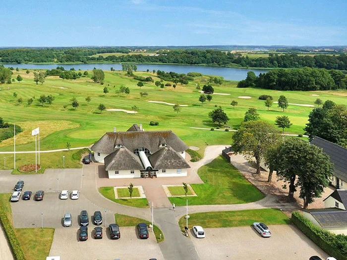 Luftfoto af en golfbane med et klubhus med stråtag, parkerede biler, grønne fairways, træer og en stor sø i baggrunden under en klar blå himmel.