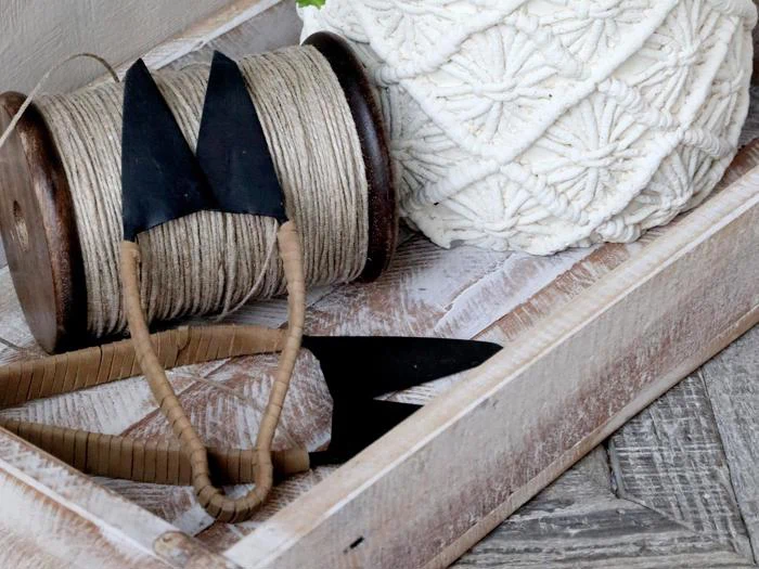 En træbakke med en rulle sejlgarn, en hvid makramékugle og to dekorative pilespidser i sort metal med omviklede håndtag.