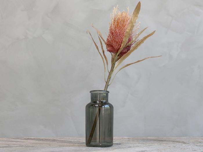 En enkelt lyserød tørret blomst med lange blade i en lille, klar grå glasvase på en træplade mod en lysegrå baggrund.