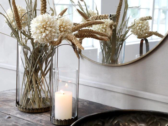 En glasvase med hvide tørrede blomster og græsser står på et træbord ved siden af et tændt stearinlys i en glasholder, og et rundt spejl reflekterer arrangementet.