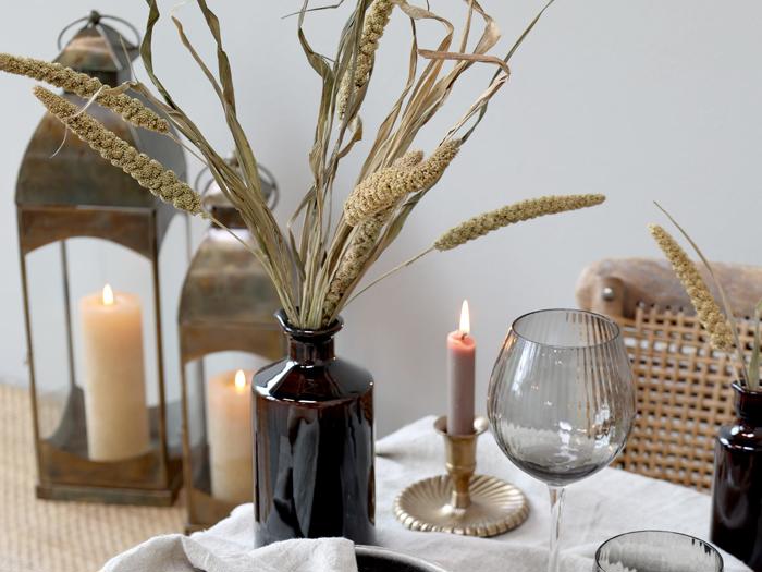Et bord med tørret græs i en mørk vase, stearinlys i metallanterner, et tændt fyrfadslys i en holder og et vinglas på en linneddug.