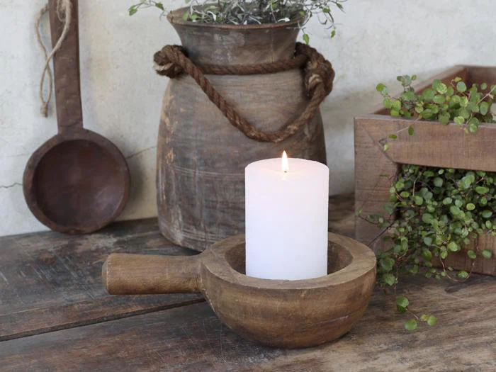 Et tændt hvidt bloklys står i en rustik træskål på et træbord, omgivet af lerkrukker og grønne planter.