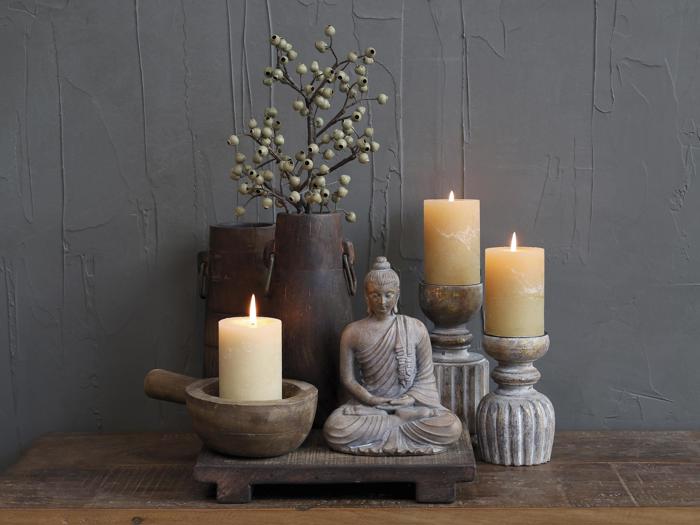 En rolig scene med en lille buddhastatue, tre tændte stearinlys, rustikke trævaser med bær og en morter og støder på en træoverflade mod en struktureret grå væg.