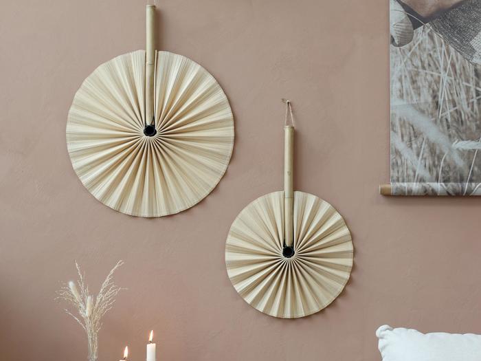 To dekorative håndvifter med bambushåndtag hænger på en lysebrun væg over en hvid pude; et tændt stearinlys og tørret græs står på et bord nedenunder.