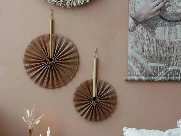 To runde, plisserede brune vifter med bambushåndtag hænger på en beige væg ved siden af et foto af en hånd, der holder en hat; stearinlys og tørrede blomster står på en nærliggende flade.