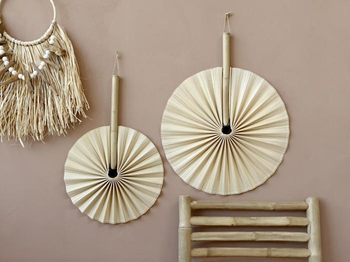 To runde, håndvævede palmevifter med bambushåndtag hænger på en beige væg ved siden af et dekorativt stykke strå og muslingeskaller; en bambusbænk står nedenunder.