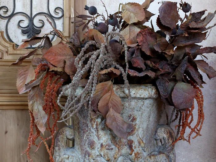 En stenkrukke rummer et arrangement af tørrede brune blade, frøkapsler og hængende orange og beige blade mod en rustik væg.