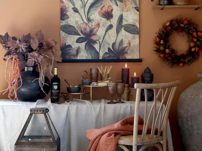Et hyggeligt bord med efterårstema med rustik indretning, stearinlys, brune glas, en krans, vin og et blomstermaleri på en orange væg, med en træstol og en hørdug.