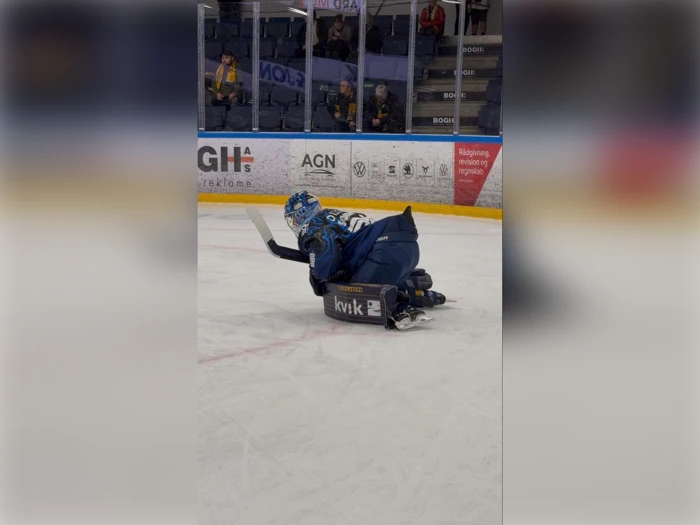 En para-ishockeyspiller i blåt udstyr ligger på sin slæde med en stav på isen under en kamp, mens der er tilskuere i baggrunden.
