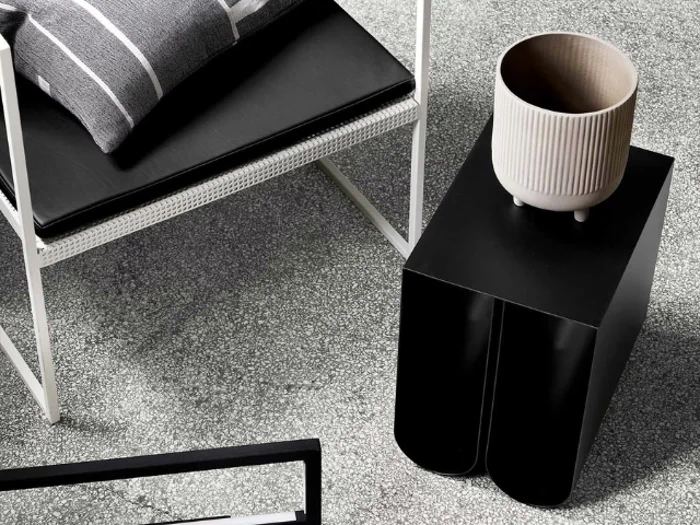 En moderne scene med en sort polstret stol, et sort sidebord og en riflet, cremefarvet krukke, alt sammen på et spættet gråt gulv.