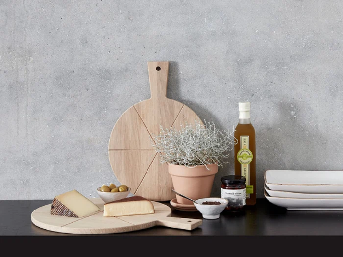 To skærebrætter af træ, en potteplante, ost, oliven, en krukke, en olieflaske, et saltfad og stablede tallerkener er pænt arrangeret på en sort flade mod en grå væg.