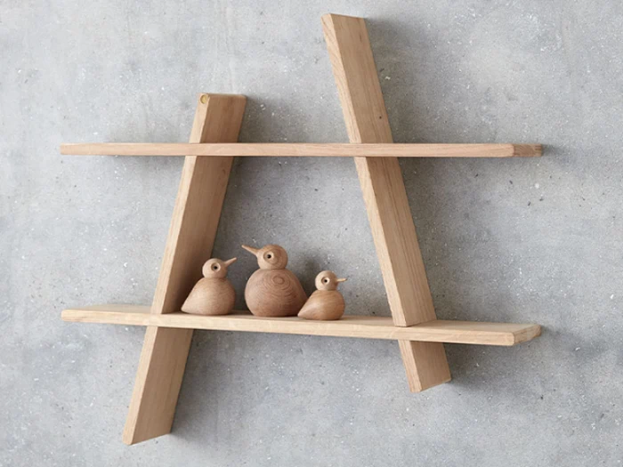 En træhylde formet som bogstavet A holder tre små fuglefigurer af træ op mod en grå betonvæg.