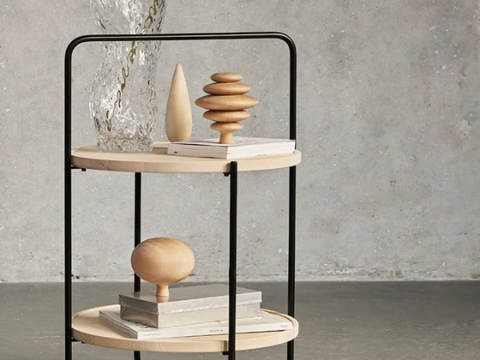 Et toetagers sidebord i træ og metal med abstrakte træskulpturer, bøger og en klar tekstureret vase på hylderne mod en grå betonvæg.