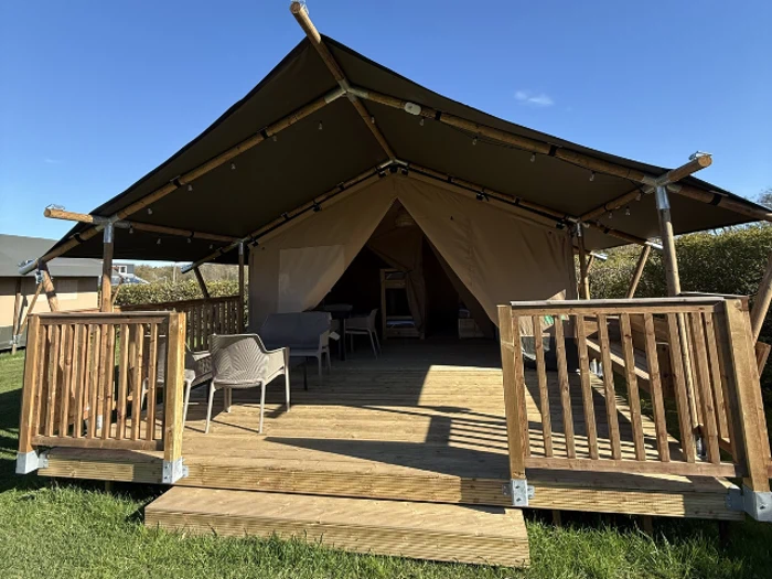 Et stort glamping-telt af lærred med en veranda af træ, udendørsstole og et bord, der står på græsset under en klarblå himmel.