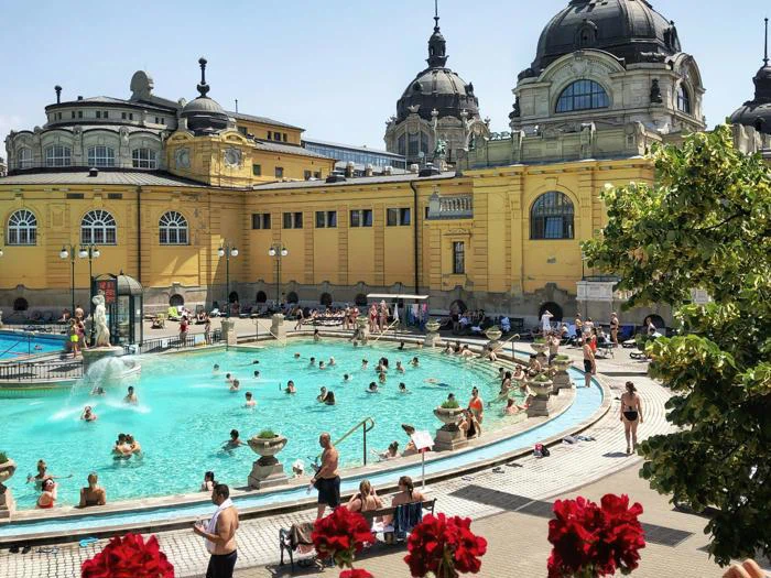 Folk svømmer og slapper af i en stor udendørs pool ved siden af en stor gul historisk bygning med kupler under en solrig himmel.
