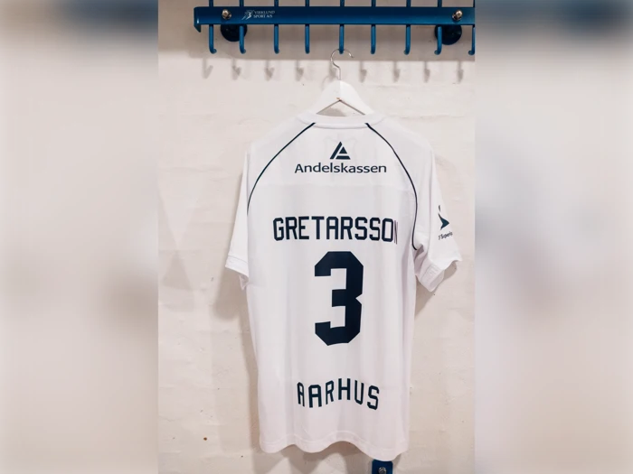 En hvid AGF Aarhus-fodboldtrøje med navnet Gretarsson, nummer 3 og et Andelskassen-logo, der hænger på et vægstativ.