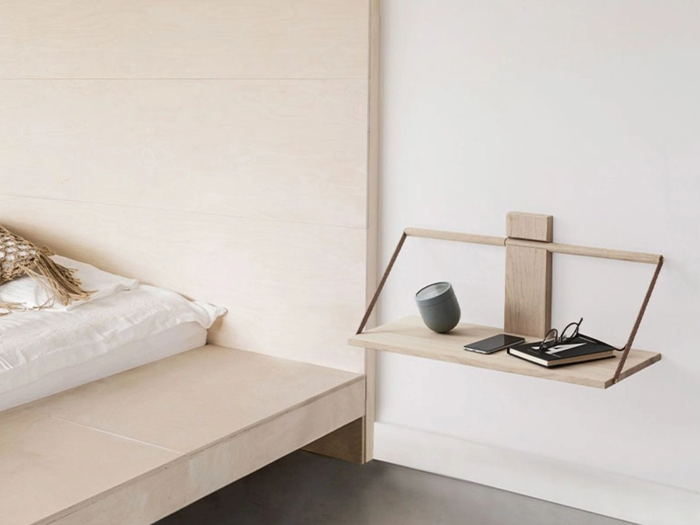 Et minimalistisk soveværelse med et svævende natbord af træ, hvorpå der står en lille højttaler, briller, en pen og en telefon, ved siden af en seng med hvidt sengetøj.