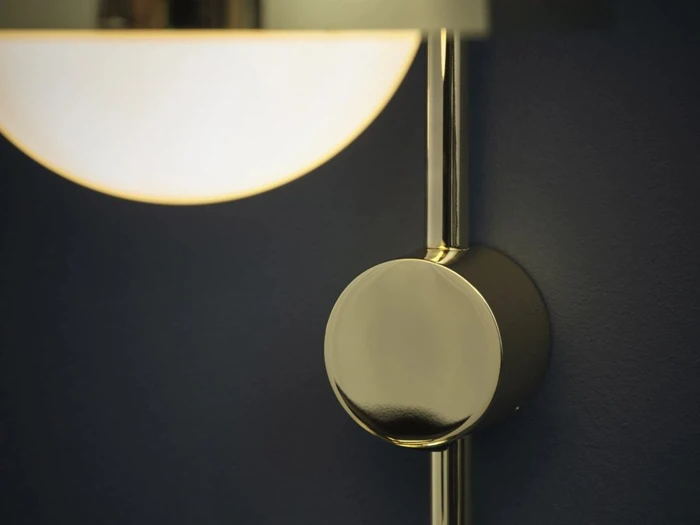 Nærbillede af en moderne væglampe med en rund armatur i messing eller guld på en mørkeblå baggrund. Lampen udsender et blødt, varmt lys.