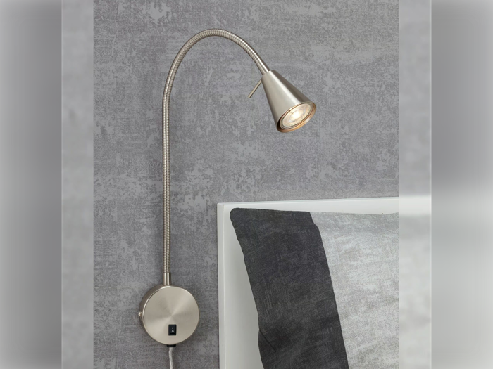 En fleksibel, metallisk vægmonteret læselampe er installeret over en seng med gråt og sort sengetøj mod en struktureret grå væg.