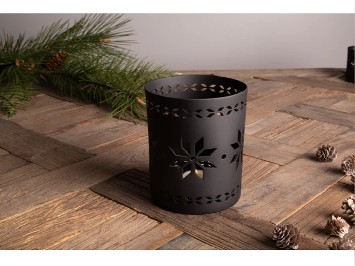 En lysestage i sort metal med udskæringer af snefnug står på et træbord ved siden af fyrretræsgrene og små kogler.