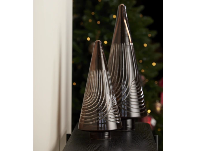 To kegleformede dekorationstræer af glas med hvirvlende mønstre står på en mørk overflade; slørede julelys og træ i baggrunden.