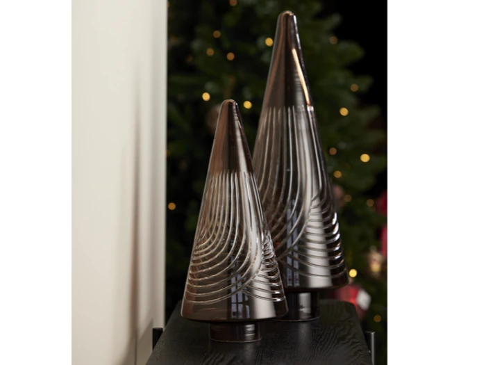To dekorative kegleformede skulpturer i mørkt glas med hvirvlende mønstre, placeret på en sort flade med et sløret, oplyst juletræ i baggrunden.