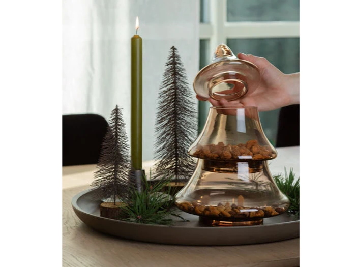 En hånd løfter låget på en glaskrukke fyldt med kæledyrsgodbidder på en bakke ved siden af et utændt grønt lys og dekorative minigraner.