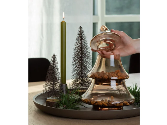 En hånd løfter låget på en glaskrukke med ravfarvede sten på en bakke ved siden af et grønt lys og dekorative miniature-fyrretræer.