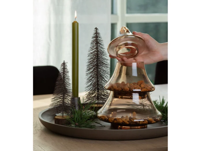 En hånd løfter låget på en glaskrukke fyldt med dyrefoder, som står på en bakke med et grønt lys og dekorative miniaturetræer.